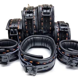 Puede incluir: Un conjunto de restricciones de cuero negro, que incluye esposas, tobilleras y un collar. Las restricciones presentan costuras decorativas multicolores y se aseguran con hebillas. Sobre un fondo blanco.