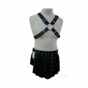 Pode incluir: Arnês de couro preto com um grande anel de metal no centro, conectado a tiras que se cruzam sobre o peito. Uma saia de couro preta combinando com tachas prateadas e uma aba lateral também está incluída. A roupa é exibida em um manequim.