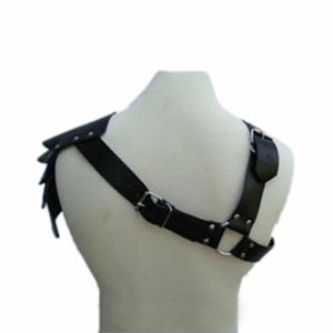 May include: Black leather shoulder armor with riveted accents and a harness design. The armor features a shoulder plate with layered, pointed edges and adjustable straps with metal buckles. This piece is designed to be worn over the shoulder.