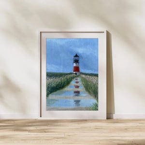 May include: Framed painting of a red and white lighthouse on a grassy hill, with a path leading to it. The sky is blue with some clouds. The artwork is in a white frame and is displayed on a wooden floor.