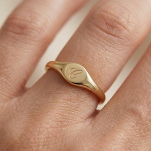 May include: A gold signet ring with a rounded rectangular face, engraved with the letter "C". The ring is worn on a finger, showcasing its polished, reflective surface. The ring's design is simple and elegant.