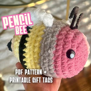 May include: A handmade, plush bee toy with a pink head, yellow and black body, and gray accents. The bee has black antennae and a large, black eye. Text on the image reads "PENCIL BEE" and "PDF PATTERN + PRINTABLE GIFT TAGS."