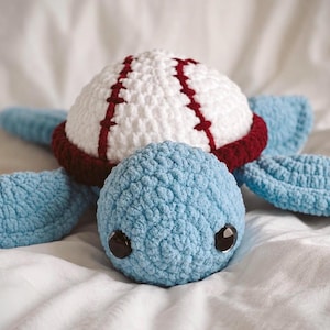 May include: A crocheted stuffed animal shaped like a sea turtle with a light blue head and flippers. The turtle shell is designed to look like a white baseball with red stitching and a dark red border around the base. It has two black plastic eyes.