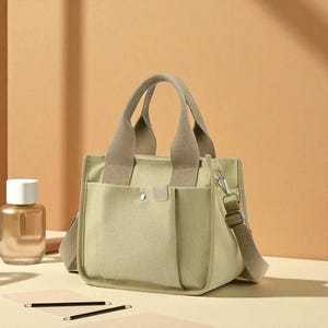 Puede incluir: Un bolso tote de lona verde claro con asa y bandolera beige. El bolso tiene dos bolsillos delanteros y un pequeño cierre de solapa. El bolso está sobre una superficie marrón claro.