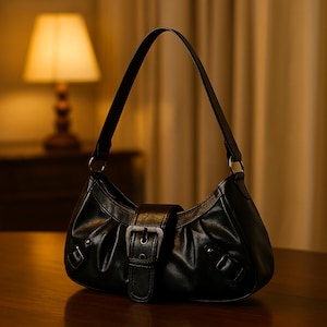 May include: Black shoulder bag with a curved design and a single strap. It has a silver buckle closure and decorative rivets. The bag is made of a smooth material, possibly leather-like.