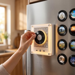 May include: A square, illuminated acrylic music player magnet featuring a central black vinyl record dial and a grid of icons. Below the dial are playback control symbols for rewind, pause, and fast forward. Several circular photo magnets are attached to the stainless steel surface nearby.