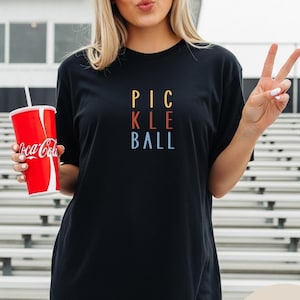 Op de afbeelding: Zwart T-shirt met de tekst "PICKLE BALL" in gele, rode en blauwe letters. Het shirt maakt deel uit van de "Comfort Colors" collectie. Een persoon houdt een rode Coca-Cola beker vast.