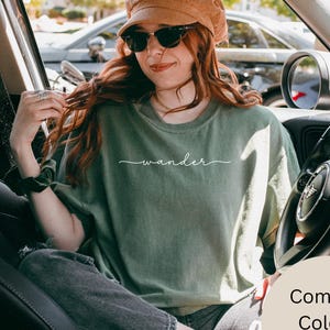 May include: An olive green t-shirt with the word "wander" in a cursive font. The shirt is part of the Comfort Colors collection and is in the color Moss. The person in the image is wearing sunglasses and a brown corduroy hat.