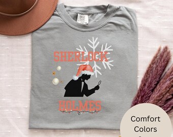 May include: A gray Comfort Colors t-shirt featuring a Sherlock Holmes design. The design includes the words "SHERLOCK HOLMES" in coral, a silhouette of Holmes wearing a Santa hat, and a snowflake. The shirt also has a string of Christmas lights.