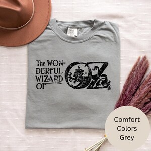 以下が含まれることがあります： グレーのTシャツ。黒の文字で「The Wonderful Wizard of Oz」と書かれています。デザインには、ライオンと文字「OZ」が入った円形のエンブレムが含まれています。シャツはComfort Colorsブランドです。