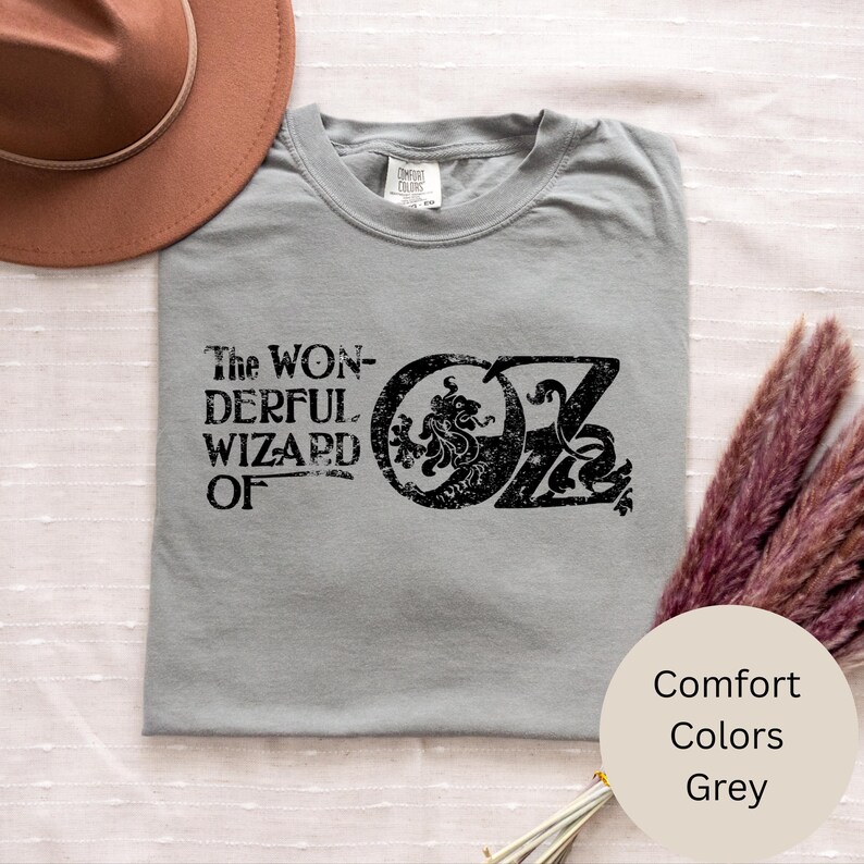 以下が含まれることがあります： グレーのTシャツ。黒の文字で「The Wonderful Wizard of Oz」と書かれています。デザインには、ライオンと文字「OZ」が入った円形のエンブレムが含まれています。シャツはComfort Colorsブランドです。