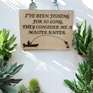 Divertente cartello da pesca – Scherzo "Master Baiter" con uomo che cattura un pesce persico – Decorazione umoristica con un pescatore – Frase di gioco di parole con un pesce persico fuori dall'acqua. File SVG digitale.