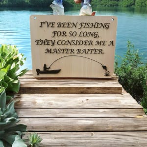 Divertente cartello da pesca – Scherzo "Master Baiter" con uomo che cattura un pesce persico – Decorazione umoristica con un pescatore – Frase di gioco di parole con un pesce persico fuori dall'acqua. File SVG digitale.