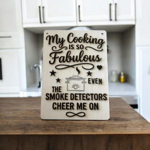 Peut inclure: Panneau rectangulaire en bois avec l'inscription "My Cooking is so Fabulous Even The Smoke Detectors Cheer Me On". Le texte noir est agrémenté d'étoiles, d'un cœur et du symbole de l'infini.