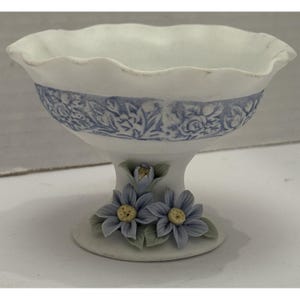 May include: A white ceramic pedestal bowl with a ruffled rim and a blue floral band. The base features two blue flower accents with yellow centers and green leaves. A decorative piece.