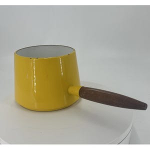 May include: A yellow saucepan with a wooden handle. The interior of the pan is white. The pan is a bright yellow color and has a long, dark brown wooden handle.