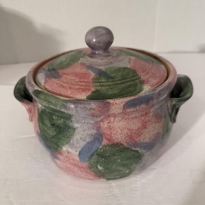 May include: A ceramic lidded pot with a round body and two handles. The pot is decorated with a pattern of pink, green, and blue splotches. The lid has a small, round knob. The pot is suitable for kitchen use.