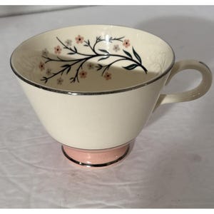 May include: Cream-colored teacup with a pink base and silver trim. The cup features a floral design with pink flowers and dark green stems. The handle is curved and the cup is on a white surface.
