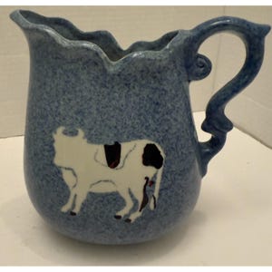 May include: A blue ceramic pitcher with a white cow design. The pitcher has a wavy rim and a curved handle. The cow is depicted in a simple, stylized manner with black spots. The pitcher is likely used for serving beverages.