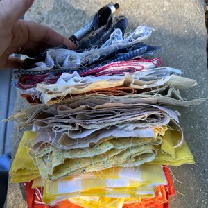 May include: A stack of assorted fabric squares featuring various colors and patterns, including yellow gingham, floral prints, solid orange, and dark blue textures. The fabric pieces have frayed, unfinished edges and are layered vertically.