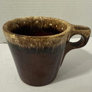 May include: A brown ceramic mug with a unique handle design. The mug features a gradient of colors, transitioning from a dark brown base to a lighter brown rim. The handle has a distinctive, open-circle design.