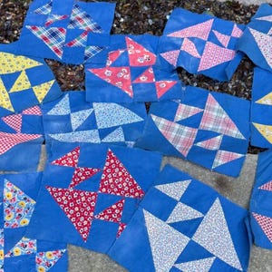 May include: A collection of square fabric quilt blocks featuring a consistent blue background with various geometric patterns in red, white, yellow, and plaid. Each block displays a unique arrangement of triangles and squares in floral and plaid prints.
