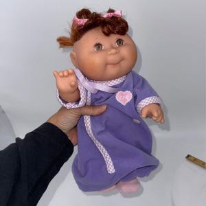 May include: A Cabbage Patch Kids doll with brown hair styled in two pigtails with pink bows. The doll has a round face, brown eyes, and is wearing a purple long-sleeved gown with a white and purple checkered trim and a heart patch labeled CPK.