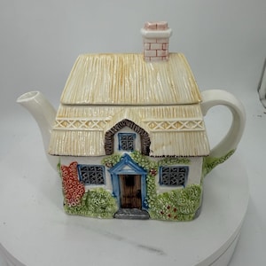 May include: A ceramic teapot shaped like a cottage with a thatched roof, a brick chimney, and a blue front door. The exterior features painted details including green bushes, red flowers, and lattice-patterned windows on a white base.