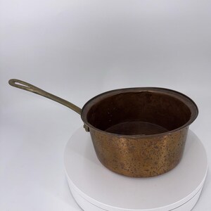 May include: A copper saucepan with a long brass handle. The pot has a rounded shape and a dark, aged patina. The handle is attached to the side of the pot with a small bracket. The saucepan is likely vintage.