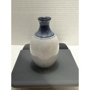 May include: A small, speckled ceramic vase with a rounded body and a narrow neck. The top and neck are a deep blue, with a lighter blue band separating them from the speckled white body. The vase is displayed on a dark gray surface.