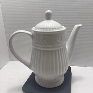 May include: A white ceramic teapot with a lid and handle. The teapot has a ribbed design on the body and decorative detailing around the top. The spout is curved, and the handle is a large, arched loop. The teapot is sitting on a dark gray surface.