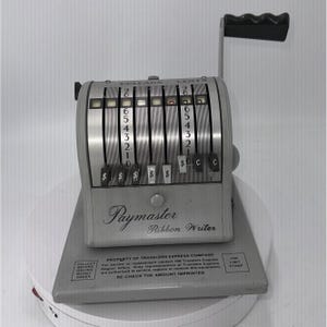 May include: A vintage Paymaster Ribbon Writer, a silver and gray mechanical device with a black handle. The machine has multiple dials for dollar and cent amounts, with the text "Paymaster Ribbon Writer" on the front.