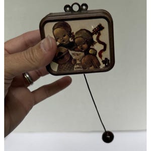 May include: A vintage, square-shaped music box with a brown wooden frame. The image inside depicts two children, one playing a stringed instrument and the other reading sheet music. A black string with a dark brown ball hangs from the bottom.