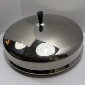 May include: A stainless steel serving dish with a domed lid and a black handle. The dish has a shiny, reflective surface and a circular base. The lid has a small, rounded black handle on top.