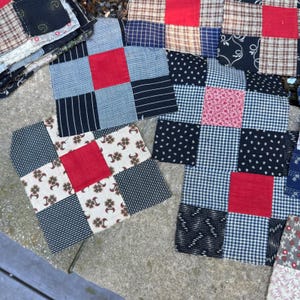 May include: A collection of square fabric quilt blocks featuring a nine-patch design. Each block displays a central red square surrounded by various patterns including black and white gingham, pinstripes, floral prints, and tan plaid fabric squares.