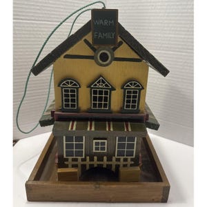 May include: A wooden birdhouse with a house-like design, featuring a yellow facade, black roof, and multiple windows. The birdhouse has a small sign that reads "WARM FAMILY" and is suspended by a green string. It sits on a wooden tray.