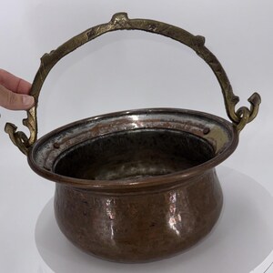 May include: A copper cauldron with a brass handle. The cauldron has a rounded body and a wide rim. The handle is arched and attached to the cauldron with decorative brass elements. The copper has a rich, aged patina.