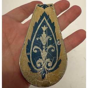 May include: A teardrop-shaped decorative object with a blue and white floral design on a gold background. The design has a cracked, aged appearance. The object is held in a hand.