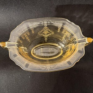 May include: A vintage, light amber-colored glass dish with a decorative gold design. The dish has an oval center and a scalloped edge with handles. The gold design includes floral and geometric patterns.