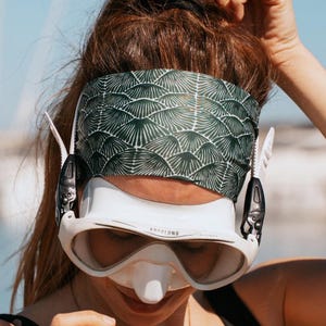 May include: A white and black diving mask with the word "Aqualung" printed on the front. The mask is worn with a green and white patterned headband. The person is wearing a black top.