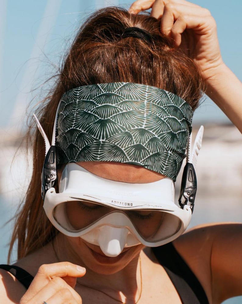 May include: A white and black diving mask with the word "Aqualung" printed on the front. The mask is worn with a green and white patterned headband. The person is wearing a black top.