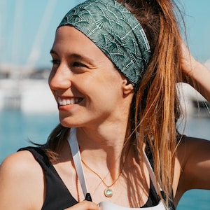May include: A green and white patterned headband is worn by a person with brown hair pulled back in a ponytail. The headband has a repeating fan-like design. A gold necklace and a black top are also visible.