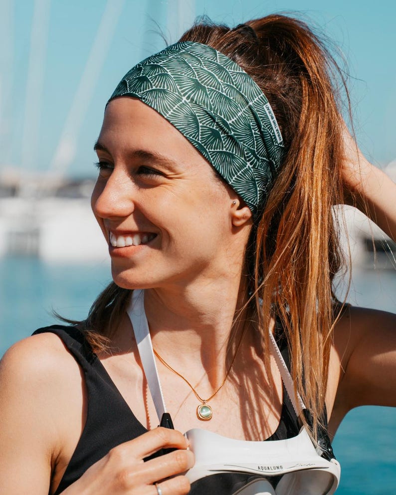 May include: A green and white patterned headband is worn by a person with brown hair pulled back in a ponytail. The headband has a repeating fan-like design. A gold necklace and a black top are also visible.