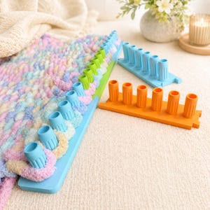 Peut inclure: Un métier à tricoter bleu clair avec une rangée de picots retenant du fil pastel panaché. Deux segments de métier supplémentaires sont présents : une pièce bleu clair à six picots et une pièce orange à six picots, toutes deux striées.