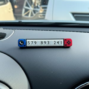 Könnte beinhalten: Eine schwarze, weiße, rote und blaue Telefonnummernanzeige für das Auto. Die Anzeige hat ein Telefonsymbol auf der linken Seite und ein rotes Knopfdessin auf der rechten Seite. Die weißen Zahlen 579 893 241 werden angezeigt.
