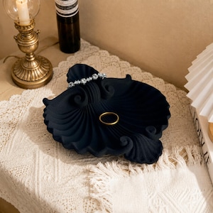May include: A matte black, shell-shaped decorative tray with a textured, ribbed surface. The tray features a silver-toned, crystal-embellished bracelet resting on its upper edge and a plain gold-toned metal ring placed in the center.