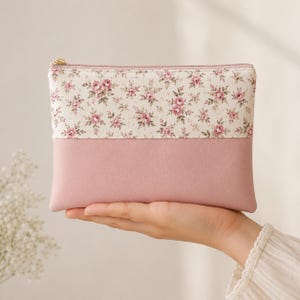 Puede incluir: Un estuche rectangular con cremallera y diseño bicolor. La sección superior es de tela color crema con un patrón repetitivo de pequeñas rosas rosadas y hojas verdes. La sección inferior es de un material liso y sólido en color rosa empolvado.