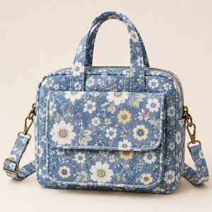 May include: A blue handbag featuring a white and yellow floral pattern. The bag has two top handles, a front flap pocket, and a detachable shoulder strap with antique brass-toned metal hardware. The main compartment closes with a zip.