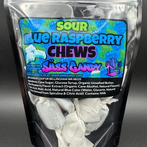 May include: A clear plastic bag filled with individually wrapped candies. The bag features a label with the words "SOUR BLUE RASPBERRY CHEWS" in large, colorful text. The label also includes the brand name "Süss Candy" and the text "Hand Crafted & Plant Based".