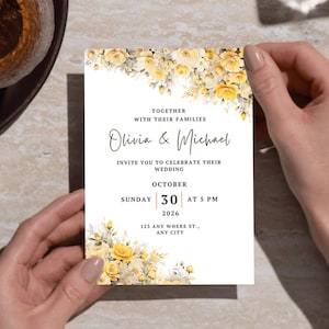 May include: A wedding invitation held by hands, featuring floral accents in yellow and white. The invitation includes the names "Olivia & Michael" and event details for October 30, 2026, at 5 PM. A glass of amber liquid and a pen are in the background.